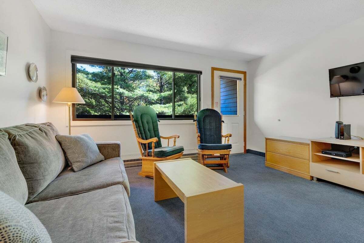 Une pièce comprenant un canapé, deux fauteuils, un téléviseur, un meuble en bois et une grande fenêtre donnant sur des arbres.