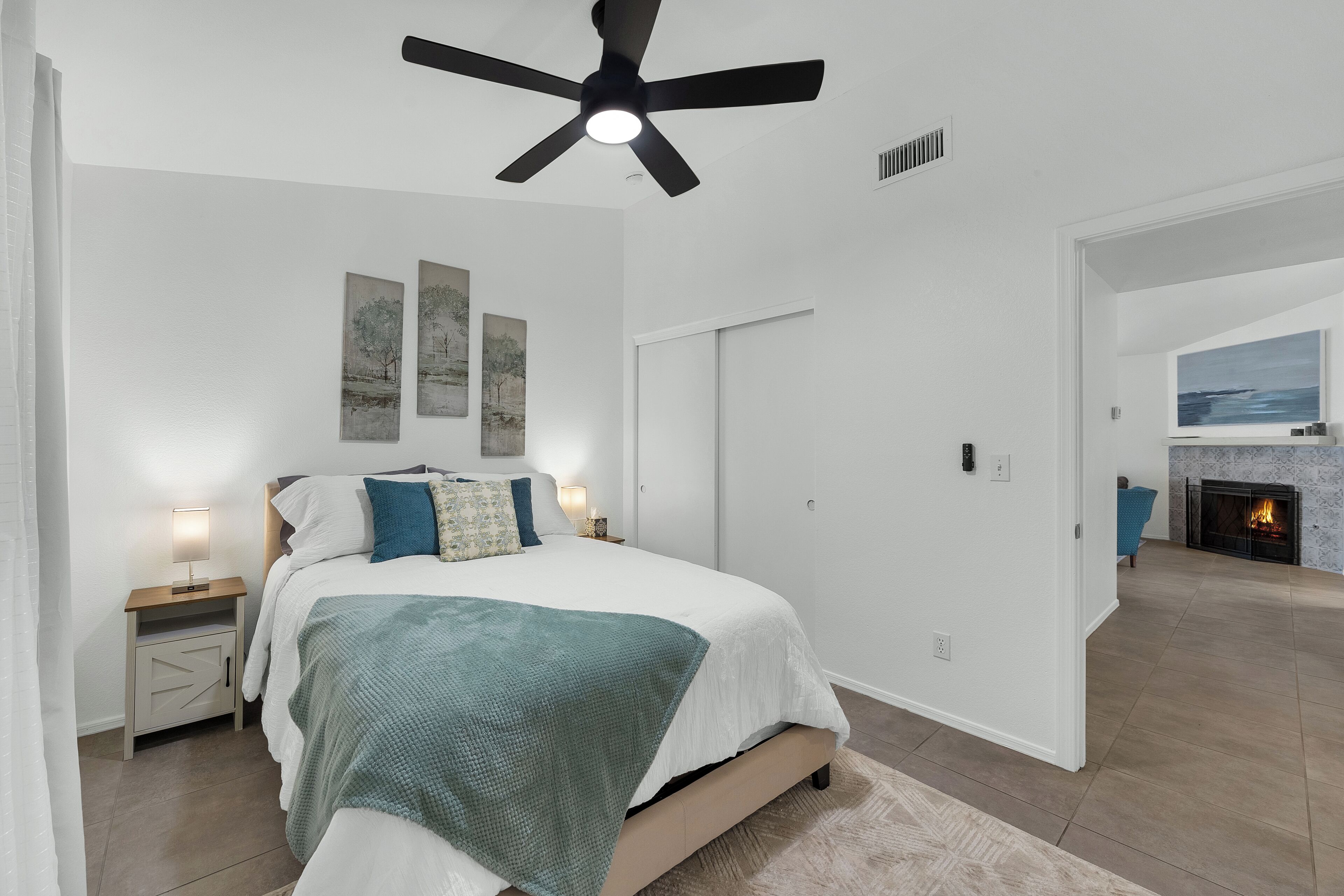 A bedroom with a bed, a ceiling fan, a nightstand, and a fireplace in the adjacent living area.