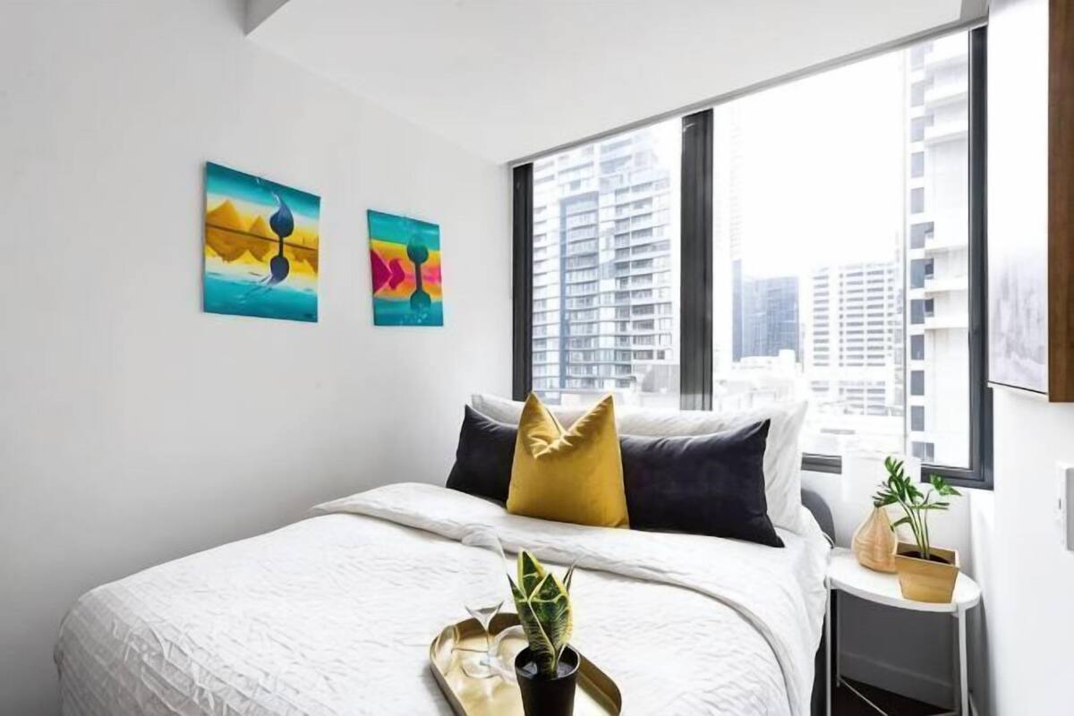 A modern bedroom with a large bed, two wall-mounted paintings, a window with a city view, and a small bedside table with a plant.