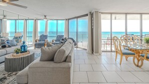 A spacious living area with a large sofa, a coffee table, and a dining table with chairs. There is a balcony with a view of the ocean.