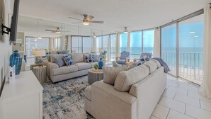 A spacious living room with a large sofa, a coffee table, and a TV. There are two ceiling fans and a large window with a view of the ocean.
