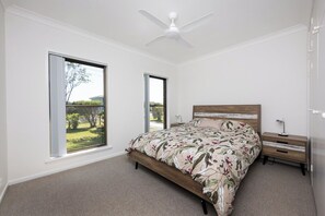 A bedroom with a bed, a nightstand, a ceiling fan, and large windows offering a view of greenery.