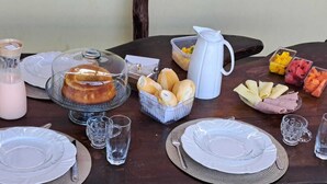 A breakfast setup with a variety of food items, including bread, fruits, and a covered cake, on a wooden table. - Pousada Sol de Berlinque (Vera Cruz)