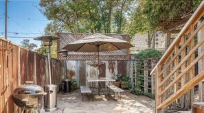 Un espacio exterior compartido con cerramiento de madera, un pérgola con sombrilla, una parrilla y un bote de basura.