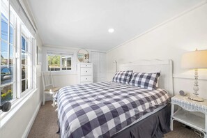 A bedroom with a checkered bedspread, a white dresser, a nightstand with a lamp, and a window with a view of the outside. - 3 Bedroom Home Near Twin Lakes Beach (Santa Cruz)