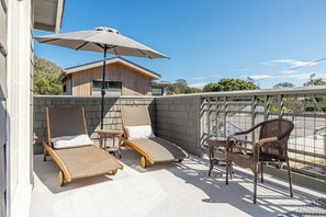 Un balcon équipé de deux chaises longues, d’un fauteuil en osier et d’un grand parasol.