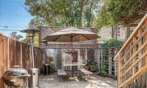 Un espacio exterior compartido con cerramiento de madera, un pérgola con sombrilla, una parrilla y un bote de basura.