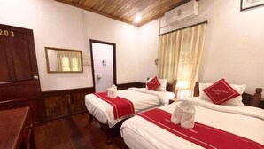 Quarto de hotel com três camas, cada uma com roupa de cama vermelha e branca, piso de madeira, porta de madeira, espelho e janela com cortinas.