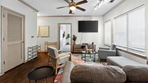 A modern living room with a ceiling fan, a flat-screen TV, and a large window with blinds.