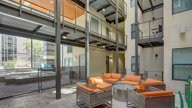 An indoor seating area with orange cushioned chairs and a round table, a metal staircase, and a view of the outside through large windows.
