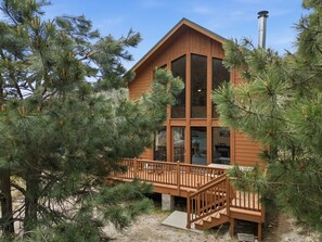 Eine zweistöckige Holzblockhütte mit großen Fenstern, einer Terrasse und umgeben von Kiefern.