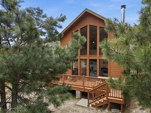 A two-story wooden cabin with large windows, a deck, and pine trees surrounding it.