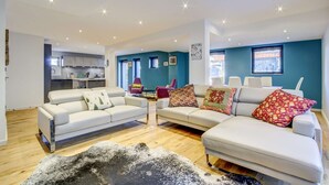 A modern living room with two white sofas, a coffee table, and a shag rug. There are two windows with curtains, a kitchen in the background, and a teal accent wall. - Very spacious apt with ground level easy access in Morzine center (MORZINE)