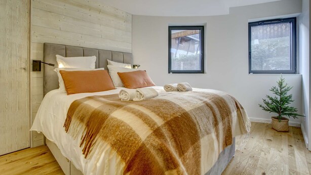 A modern bedroom with a wooden headboard, a large bed with a striped blanket, two windows, and a small potted plant. - Very spacious apt with ground level easy access in Morzine center (MORZINE)