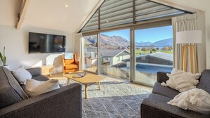 Ein modernes Wohnzimmer mit einem großen Glasfenster zum Balkon, einem Flachbildfernseher, einem Sofa, einem Sessel und einer Lampe.