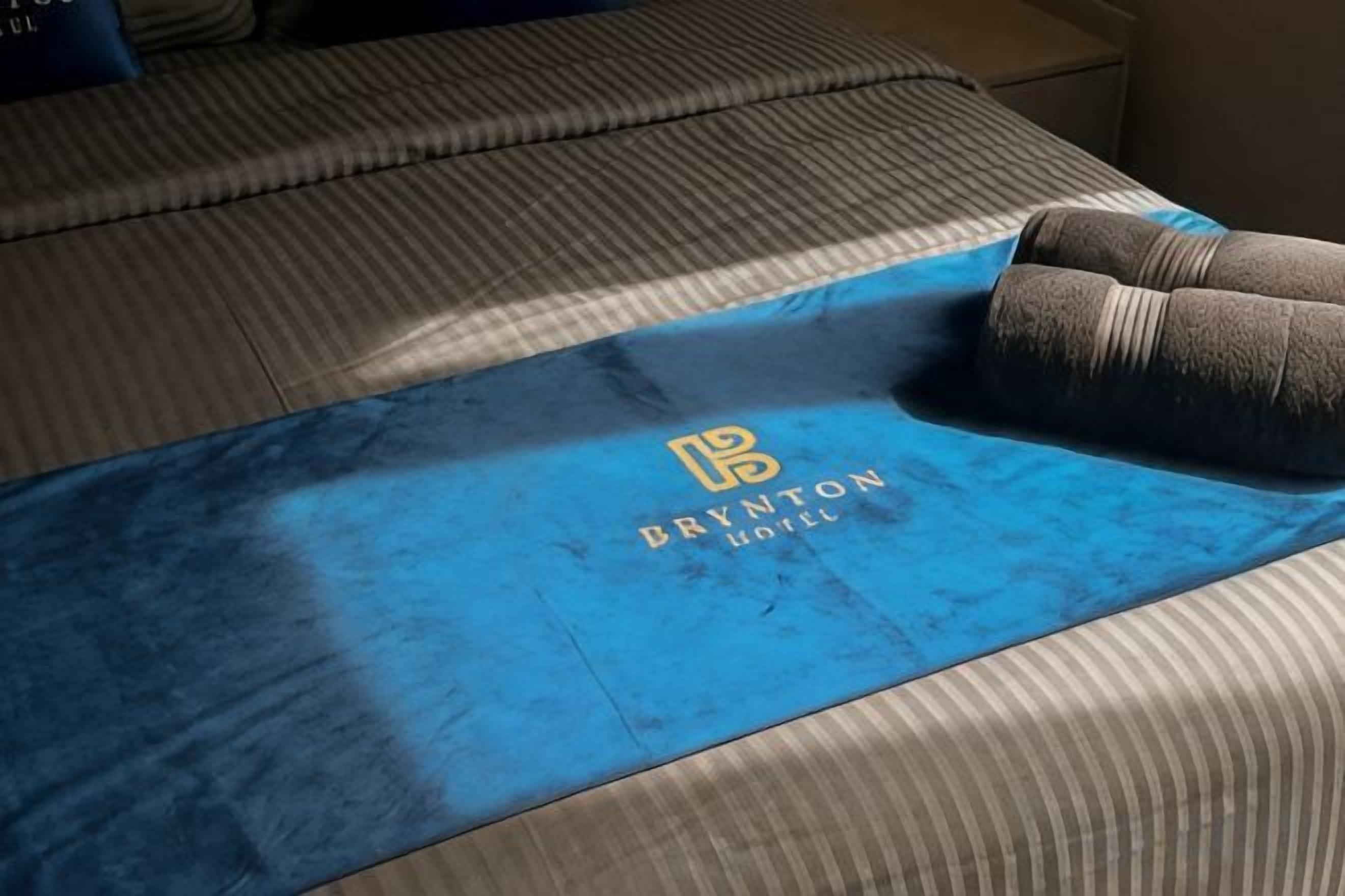 A bed with a blue blanket featuring the Brynton Hotel logo.