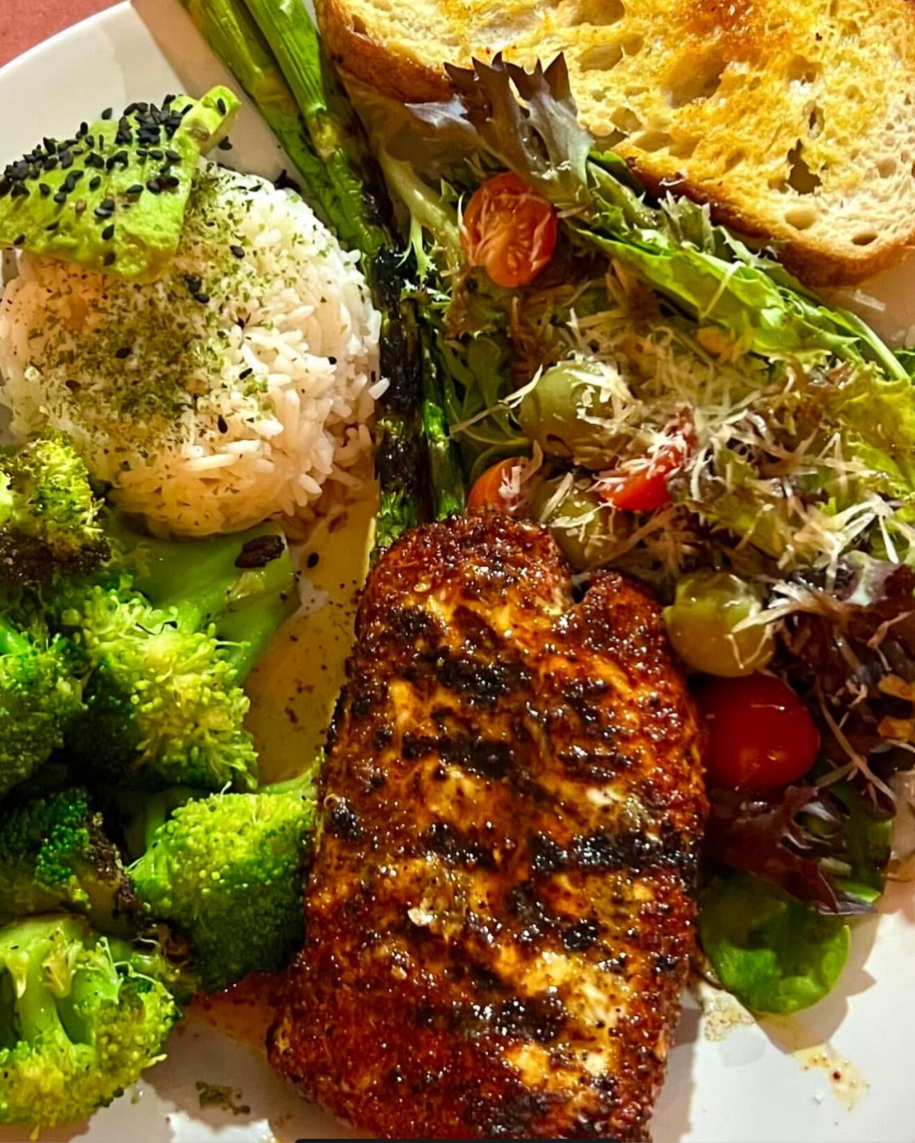 Un plato con pollo a la parrilla, brócoli, arroz y una ensalada de lechugas variadas.