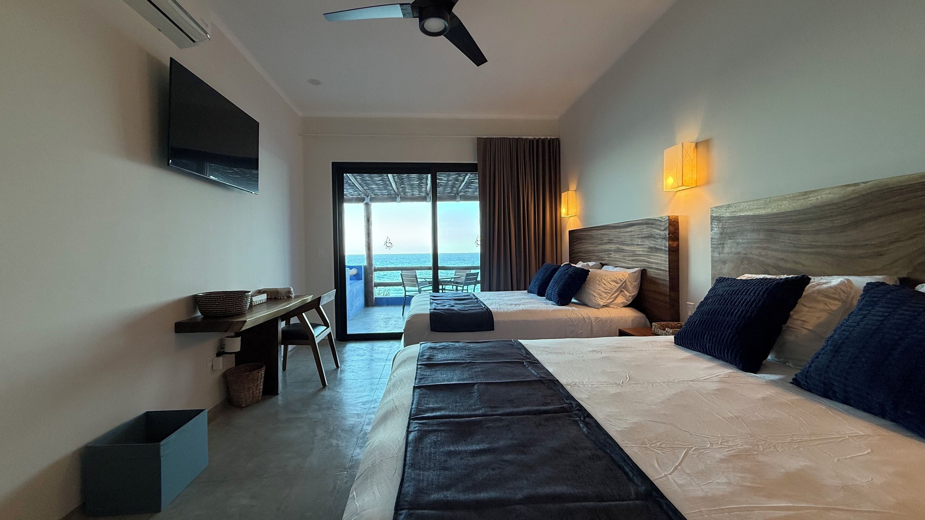 Habitación de hotel con dos camas, un escritorio, televisor y balcón con vista al mar.