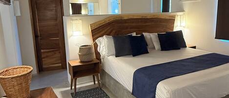 A bedroom with a wooden headboard, a bed with white and dark blue bedding, a wooden nightstand with a lamp, a woven basket, and a wooden bench.