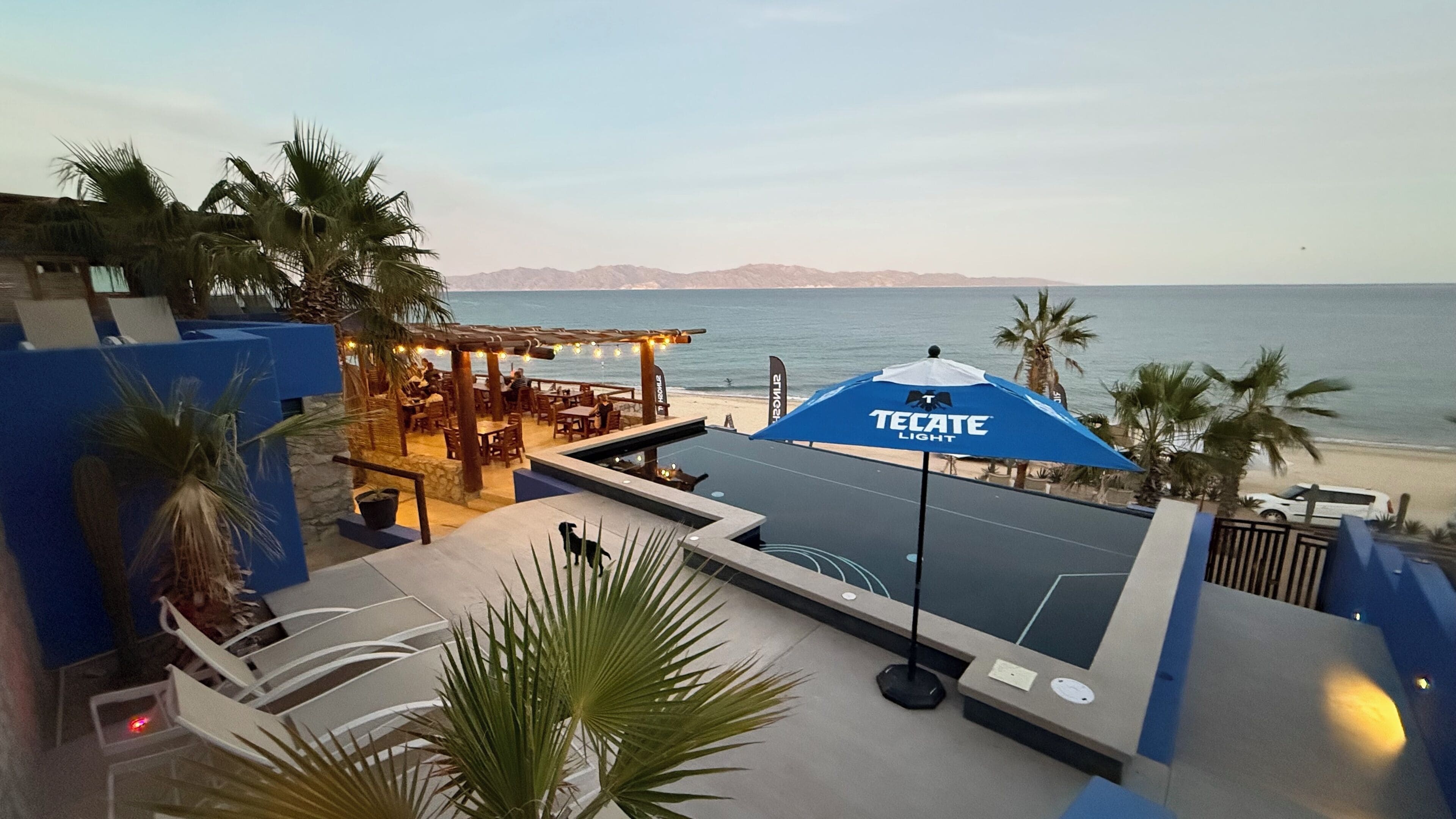 An outdoor seating area with a blue umbrella labeled "TECATE LIGHT," overlooking a beach with palm trees and mountains in the distance.