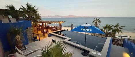 An outdoor seating area with a blue umbrella labeled "TECATE LIGHT," overlooking a beach with palm trees and mountains in the distance.