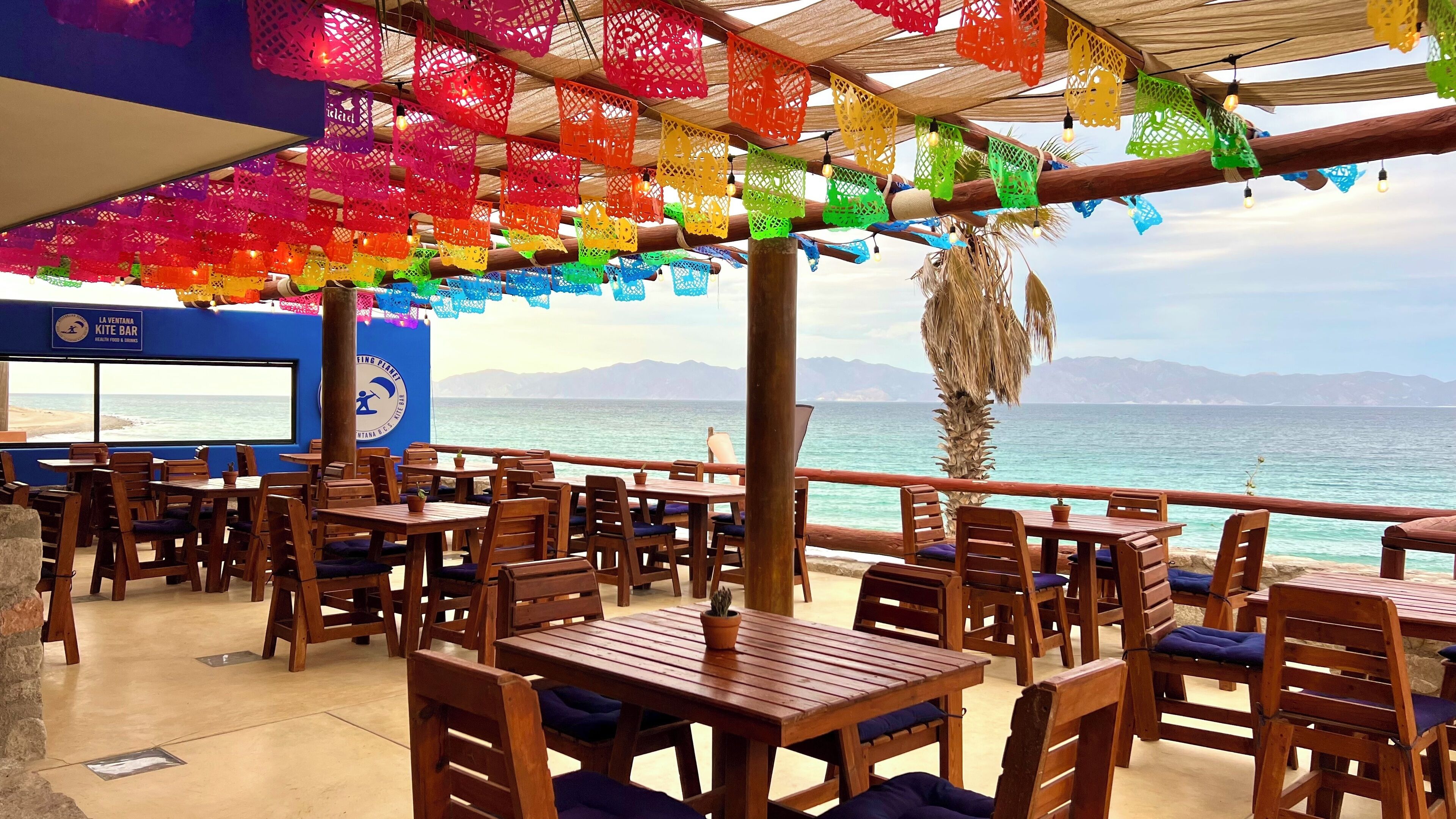 An outdoor seating area with wooden tables and chairs, a view of the sea, and colorful paper lanterns hanging from the ceiling.