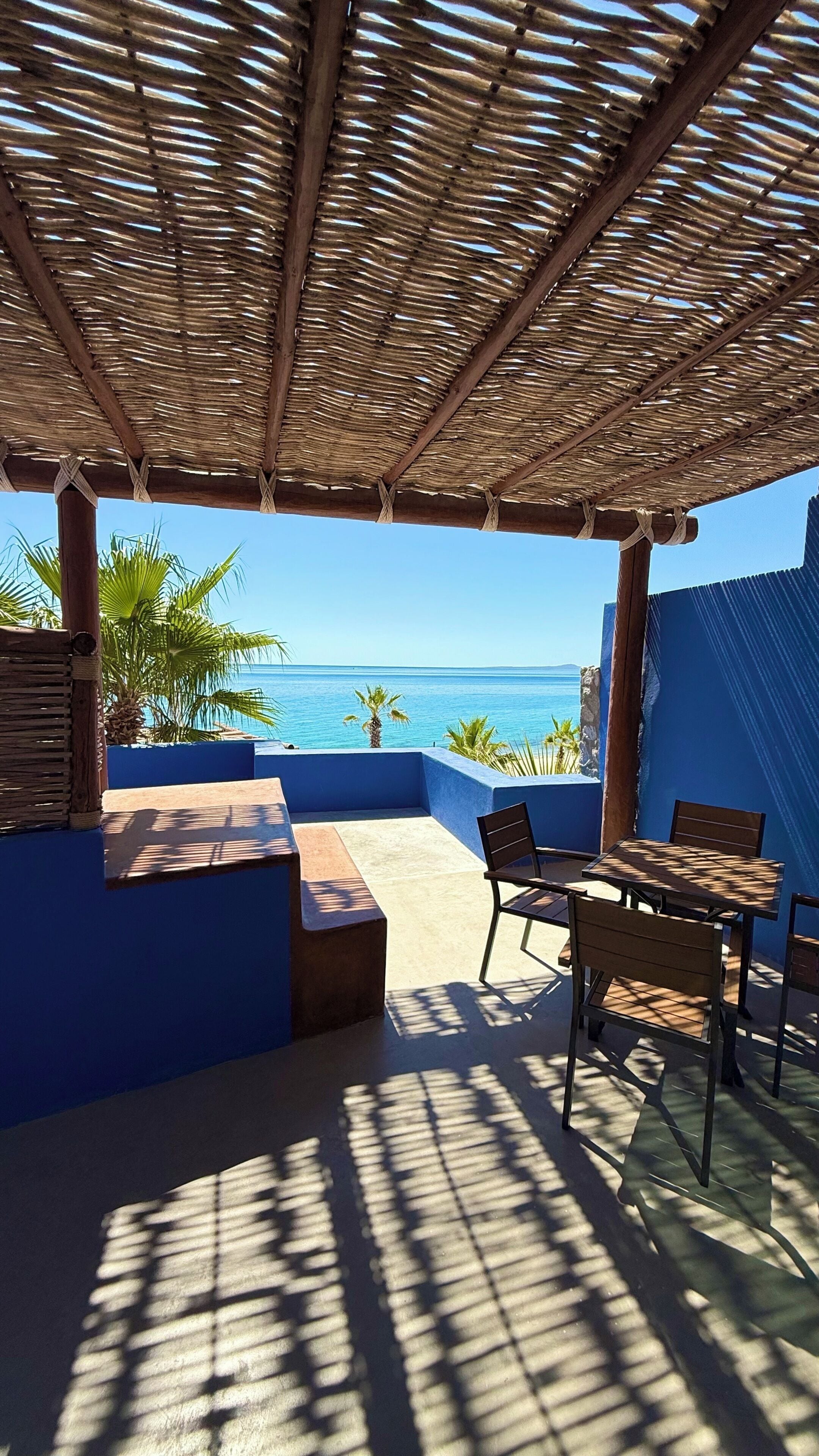 A covered outdoor seating area with a view of the sea, featuring a thatched roof, blue walls, and wooden furniture.