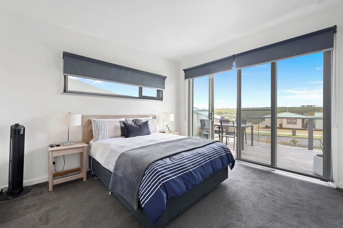 A bedroom with a large bed, a bedside table with a lamp, a heater, and a view of the outdoors through a sliding glass door.
