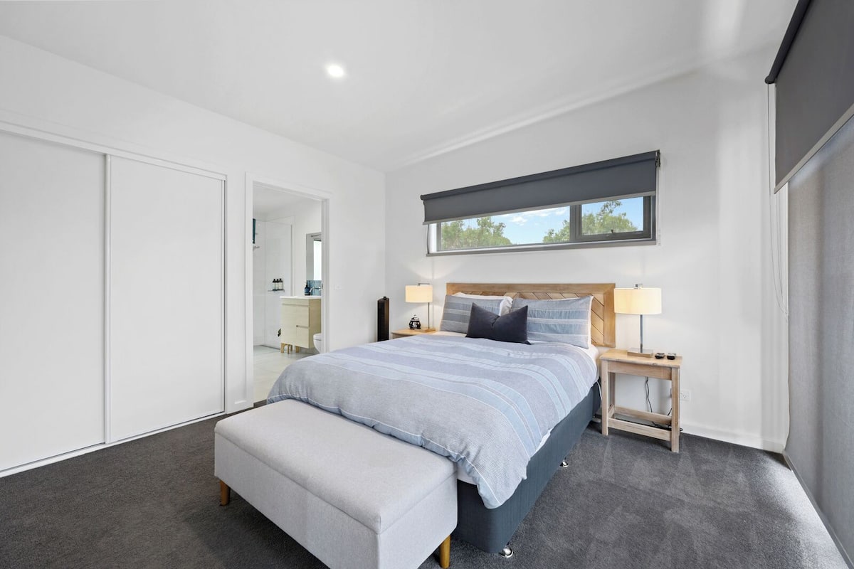 A modern bedroom with a large bed, bedside tables, a bench, and a view of the outdoors through a window.