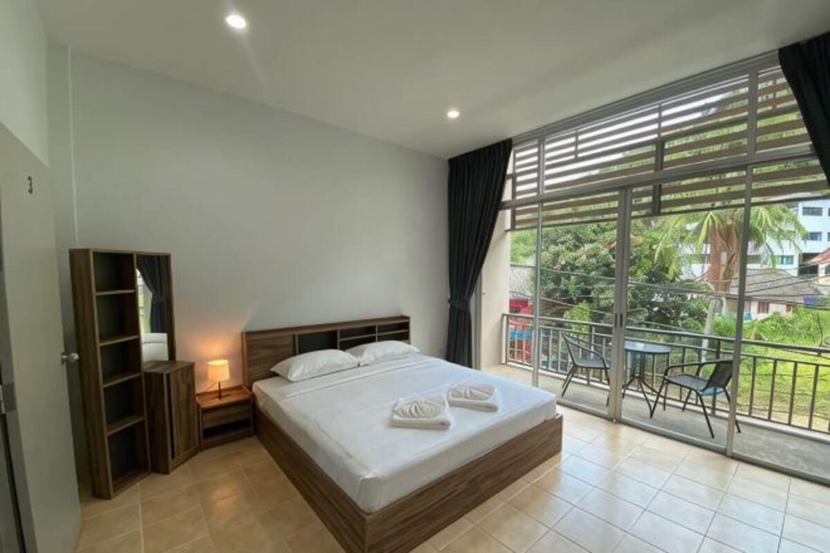 A bedroom with a large bed, a nightstand, a wardrobe, and a balcony with a view of greenery and buildings.