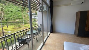 A hotel room with a balcony, a bed, a table with chairs, and a view of greenery and buildings outside. - L&P Guesthouse (Karon)