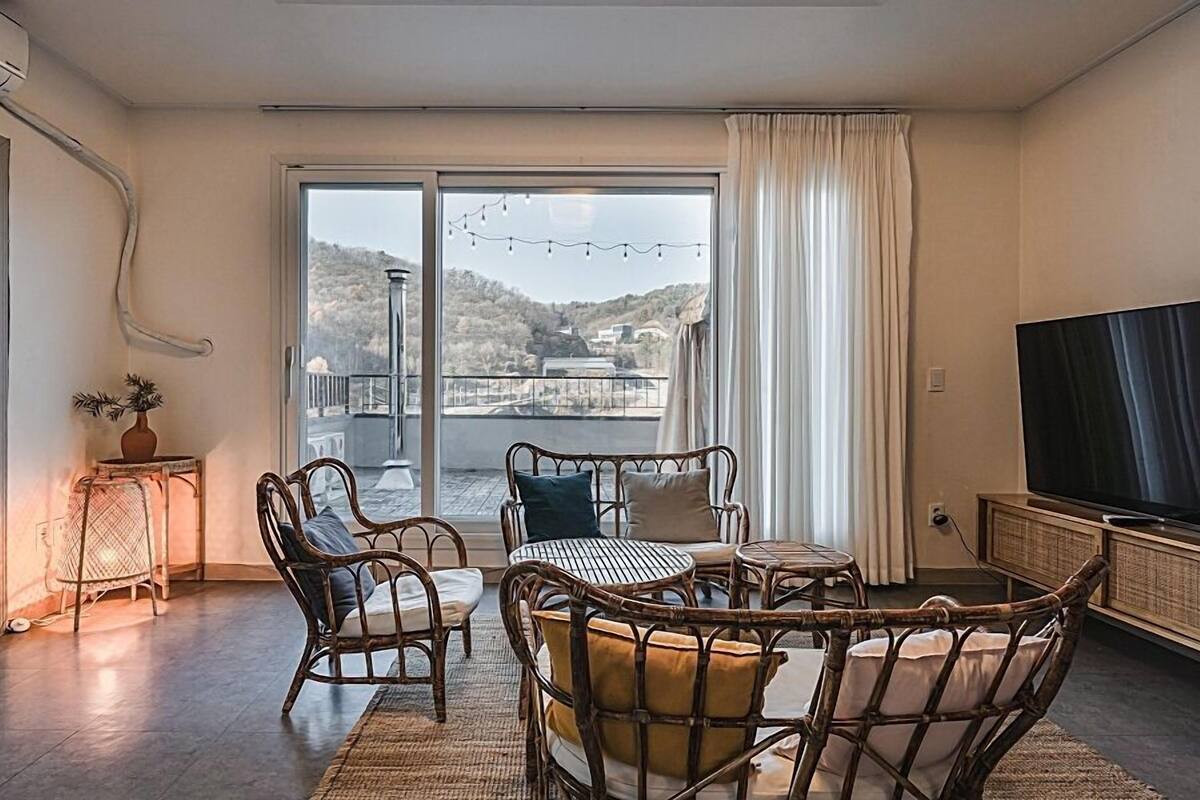 A cozy room with a balcony, wicker furniture, a television, and a view of a hilly landscape.