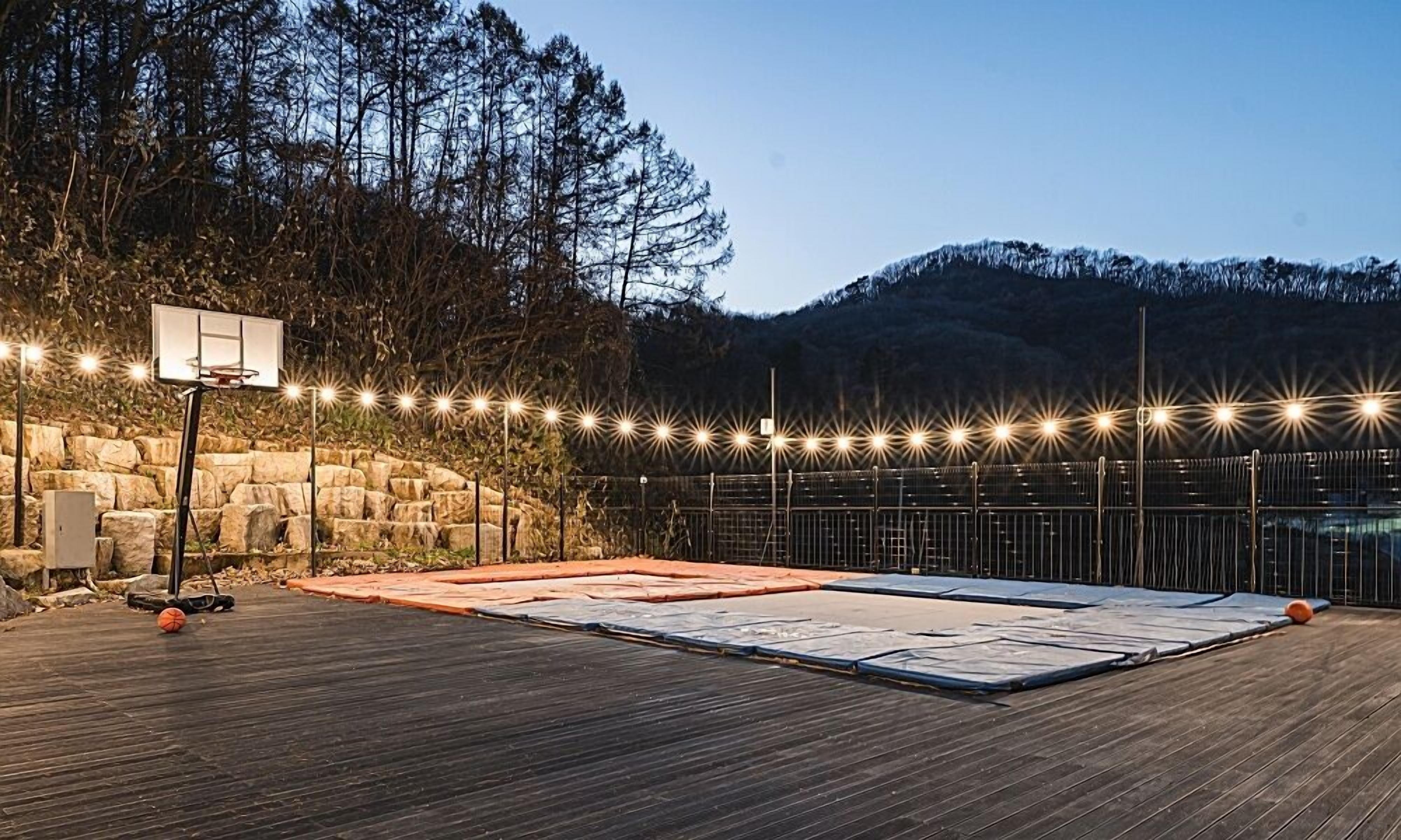 An outdoor basketball court with a wooden floor, a basketball hoop, and a string of lights.
