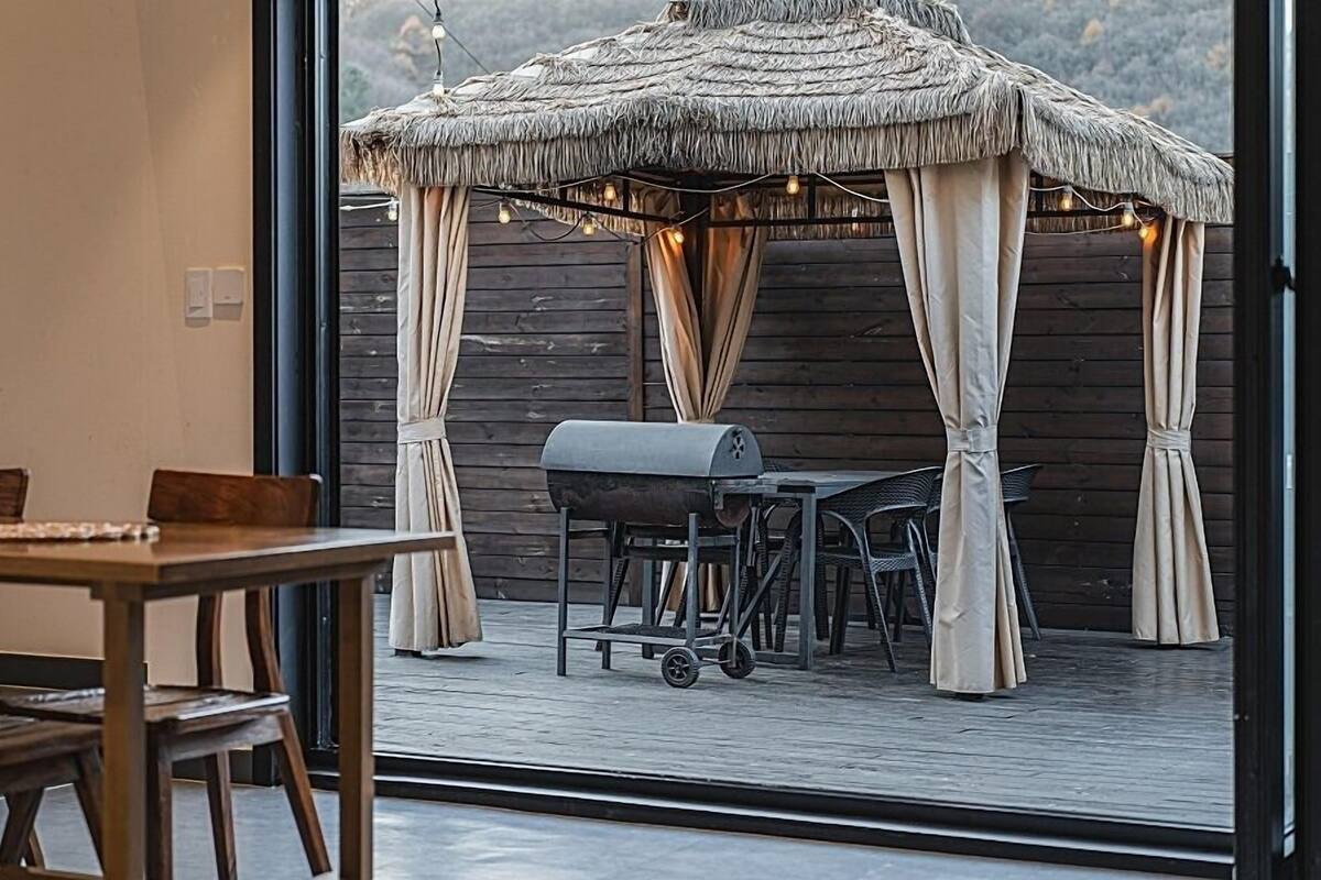 An outdoor dining area with a wooden deck, a grill, and a thatched gazebo.