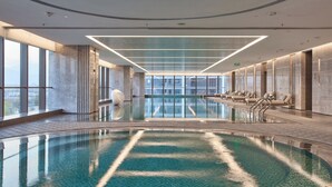 A modern indoor swimming pool with clear water, a lounge area with chairs, and large windows offering a city view. - Shilla Monogram Xi'an (Xi'an)