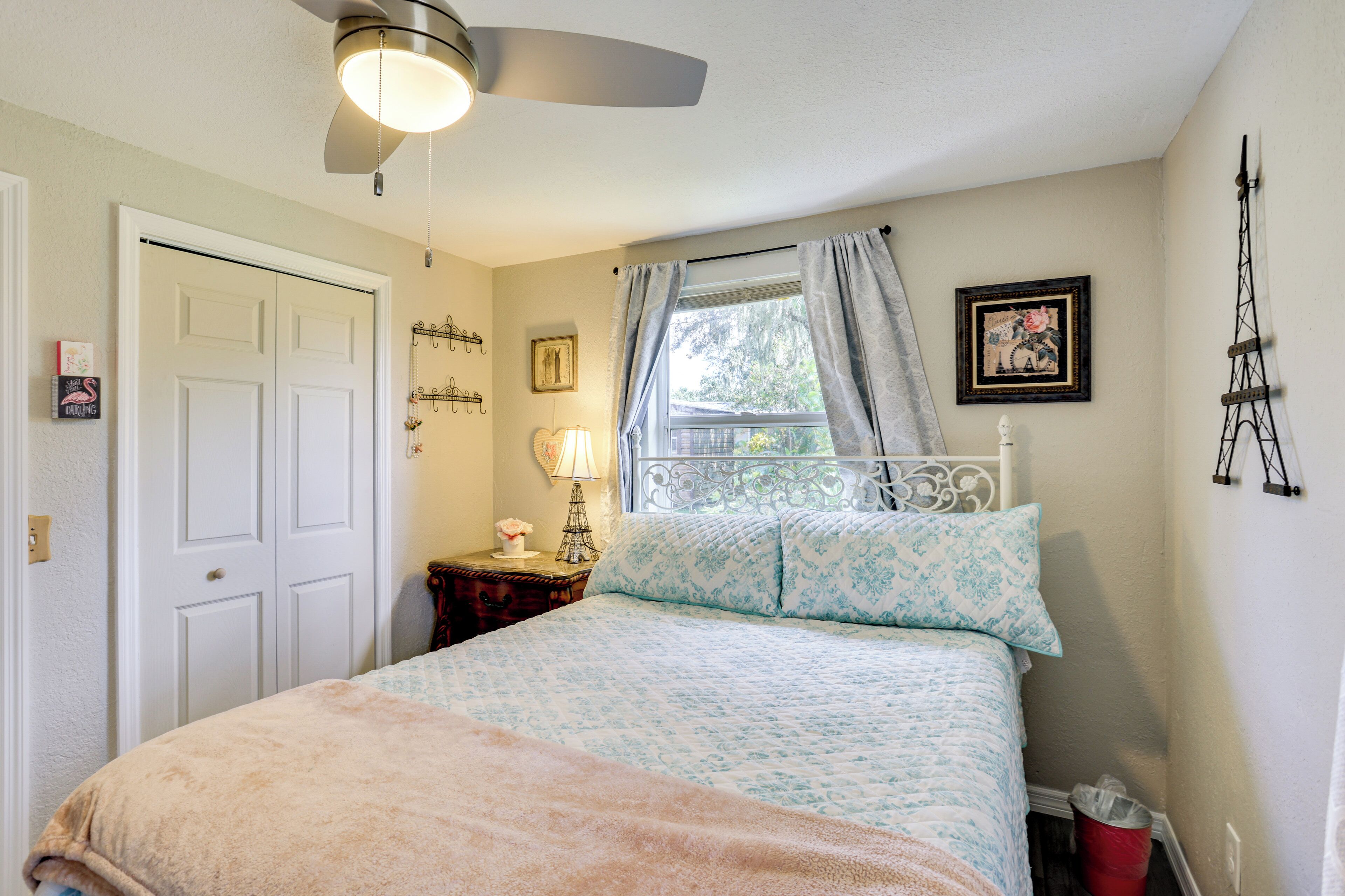 A bedroom with a bed, a ceiling fan, a window with curtains, a nightstand with a lamp, and a wall-mounted picture.