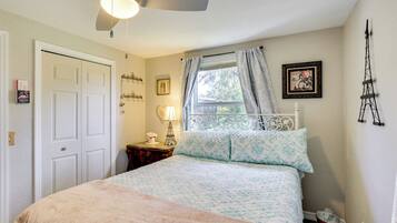 A bedroom with a bed, a ceiling fan, a window with curtains, a nightstand with a lamp, and a wall-mounted picture.