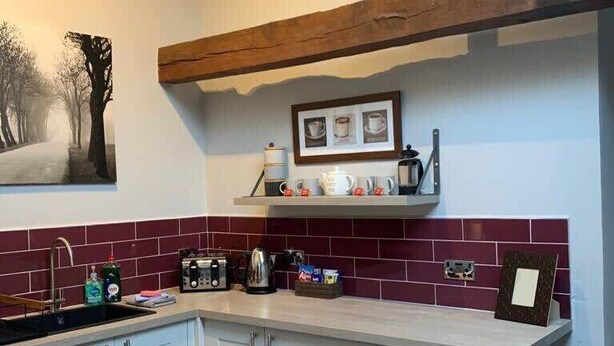 Une cuisine moderne avec des armoires blanches, un plafond avec une poutre en bois, un dosseret de carreaux rouges et un tableau encadré au-dessus de l’évier.