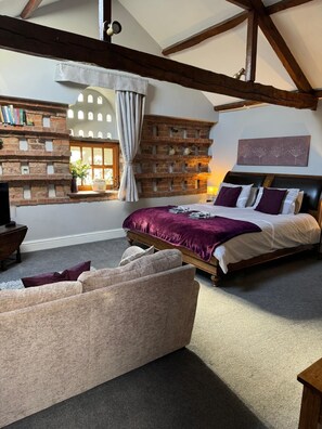 A bedroom with exposed wooden beams, a brick accent wall, a bed with a purple bedspread, and a window with curtains. - Studio 3B, The Carriage House. (Bilbrough)