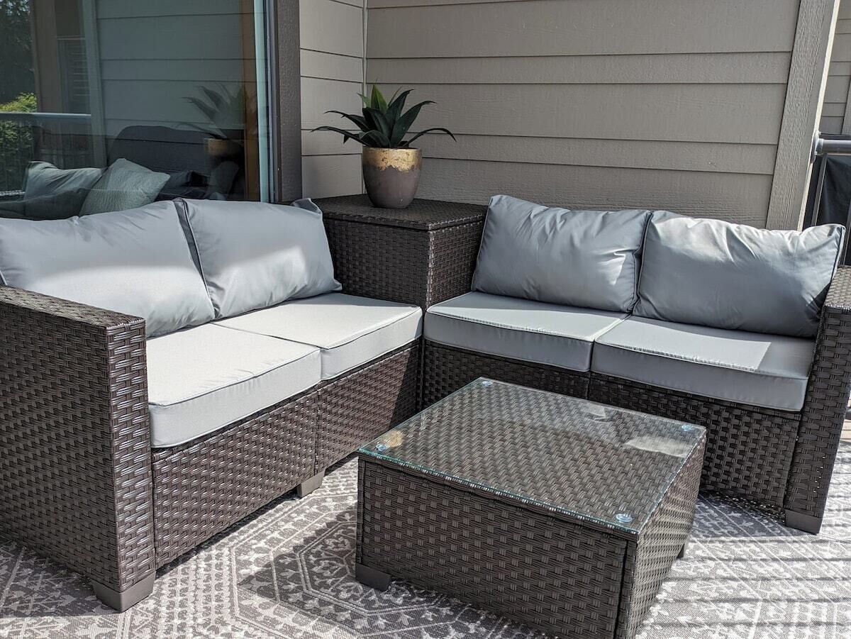 Un canapé d’angle en osier avec des coussins gris et une table basse en verre, le tout installé dans un espace extérieur.