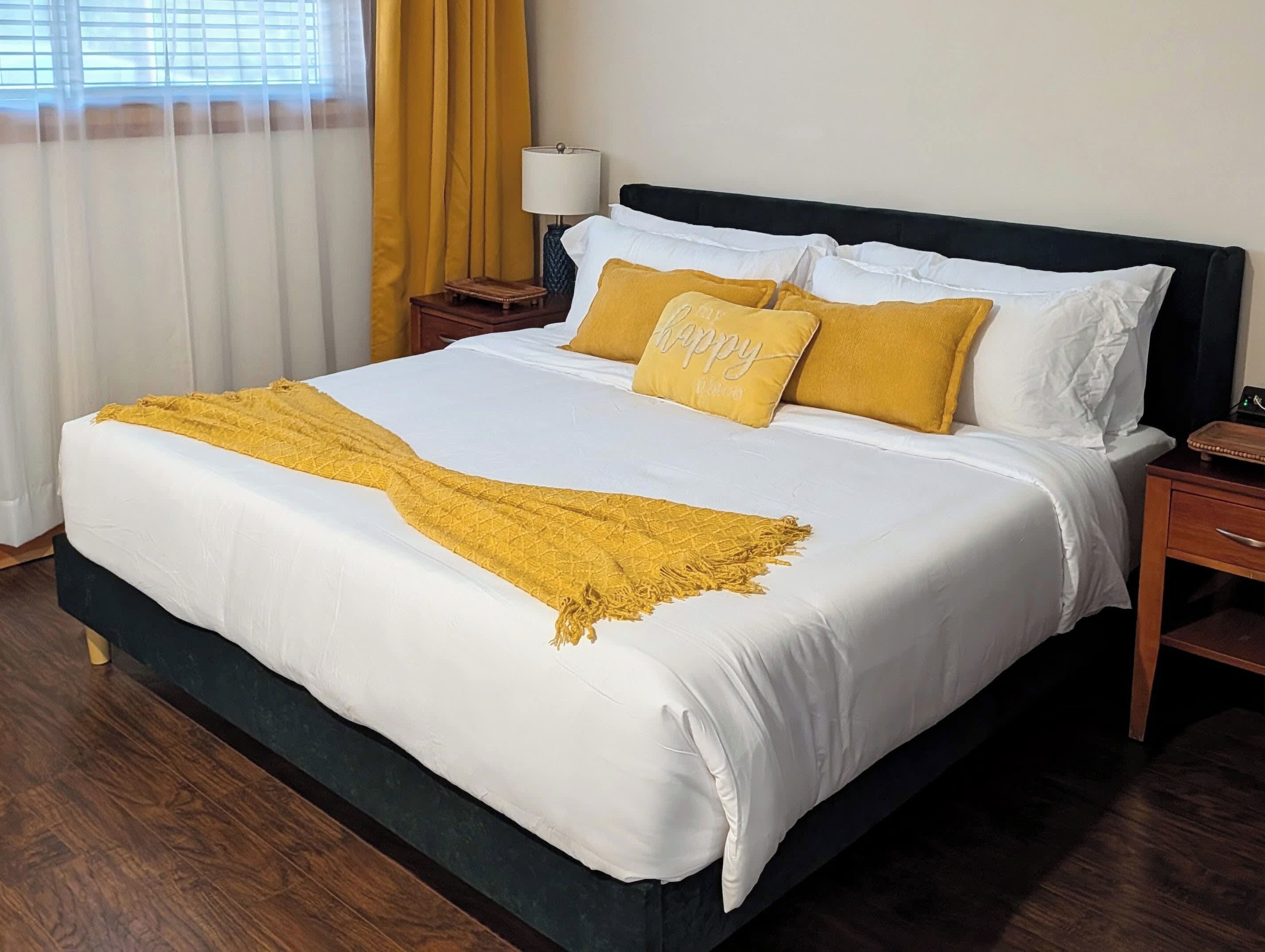 Een netjes opgemaakt bed met witte beddengoed en een geel plaid, een nachtkastje met een lamp, en een raam met gele gordijnen.