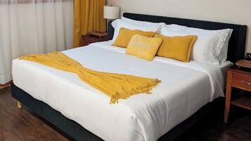 Een netjes opgemaakt bed met witte beddengoed en een geel plaid, een nachtkastje met een lamp, en een raam met gele gordijnen.