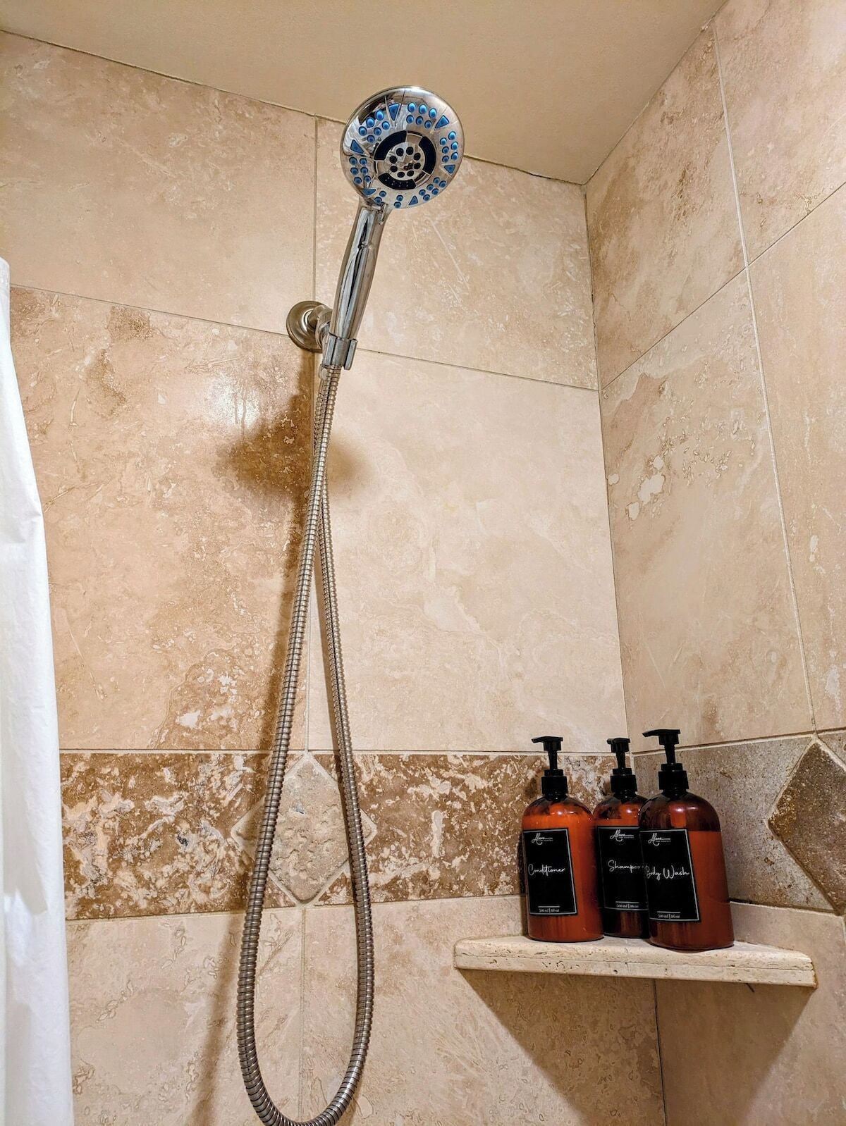 A shower with a handheld showerhead and a flexible hose. There are three bottles of shampoo and conditioner on a shelf.