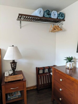 Une chambre avec une table de chevet en bois, une lampe, une petite étagère avec des serviettes pliées, une chaise en bois et une plante en pot.