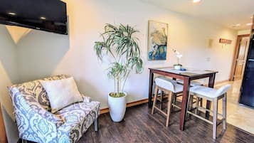 A living room with a sofa, a dining table with chairs, a potted plant, and a flat-screen TV mounted on the wall.