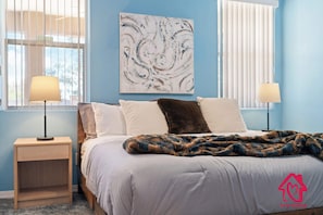 A bedroom with a bed, two bedside lamps, a blue wall, a large abstract painting, and a logo on the bedspread. - Rancho de los Sueños with Hot Tub - An Irvie Home (Albuquerque)