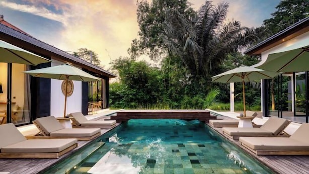 Piscina con sillas de descanso y sombrillas, rodeada de exuberante vegetación y arquitectura moderna.
