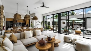 Una sala de estar moderna con sofá, mesa de centro y sillones, ventanas amplias con vistas al exterior y una zona de cocina.