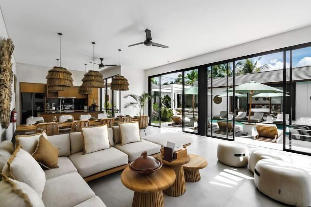 Un salon moderne comprenant un canapé, une table basse et des fauteuils, de grandes fenêtres offrant une vue sur l’extérieur, ainsi qu’un coin cuisine.