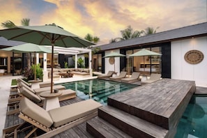 A pool area with lounge chairs and umbrellas, a wooden deck, and a modern building with a circular wall decoration. - Villa Cocktail and Dreams by BaliSuperHost (Bali)